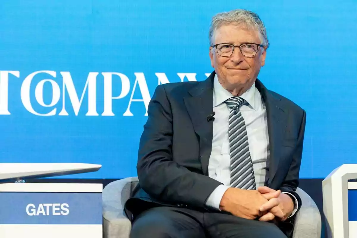 Bill gates assegut en un escenari amb fons blau i vestit amb un vestit fosc i corbata de ratlles