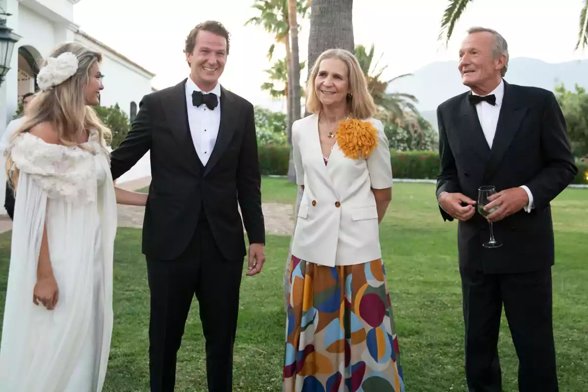 Cristina Serrano, Luis Astolfi Jr, la infanta Elena i Luis Astolfi elegantment vestits posen i somriuen a l’aire lliure en un jardí, amb palmeres i muntanyes al fons.
