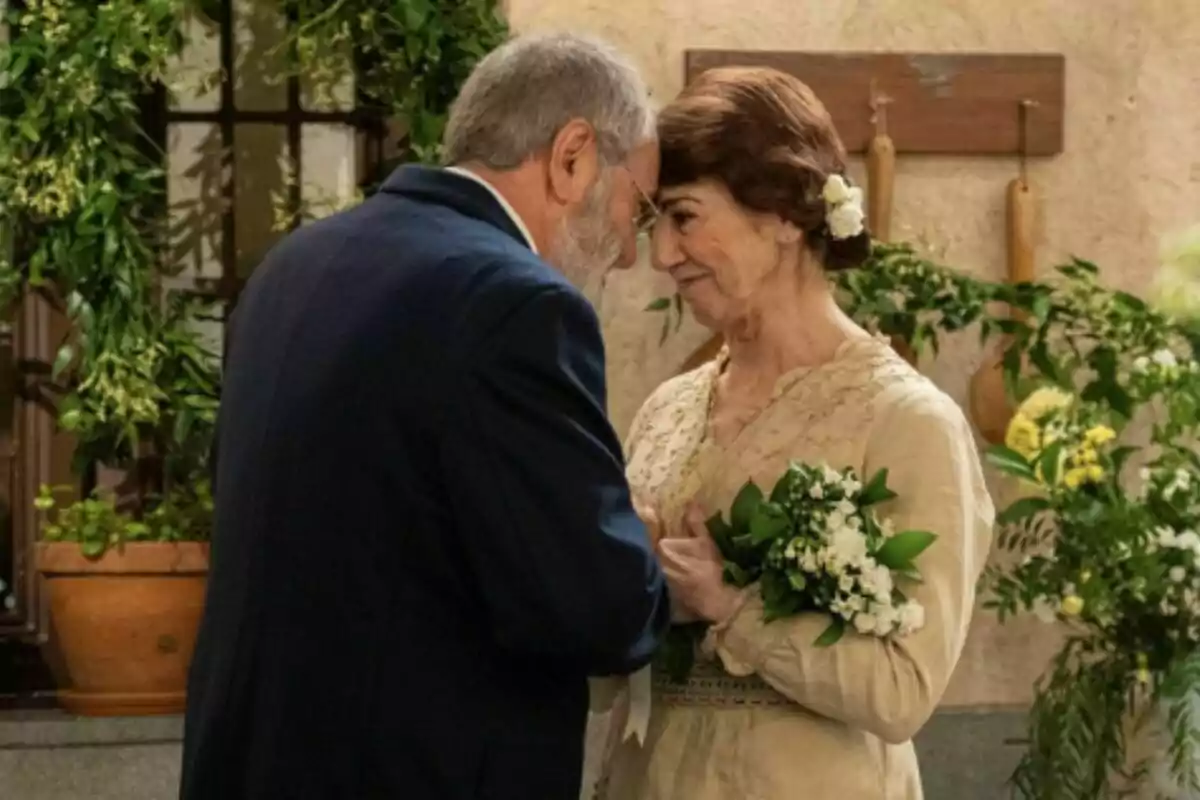 Parella de persones grans, formada per Ròmul i Emília a 'La Promesa', vestides de manera elegant es miren amb tendresa i es toquen el front mentre la dona sosté un ram de flors blanques envoltats de plantes i decoració natural. Parella de persones grans, formada per Ròmul i Emília a 'La Promesa', vestides de manera elegant es miren amb tendresa i es toquen el front mentre la dona sosté un ram de flors blanques envoltats de plantes i decoració natural.