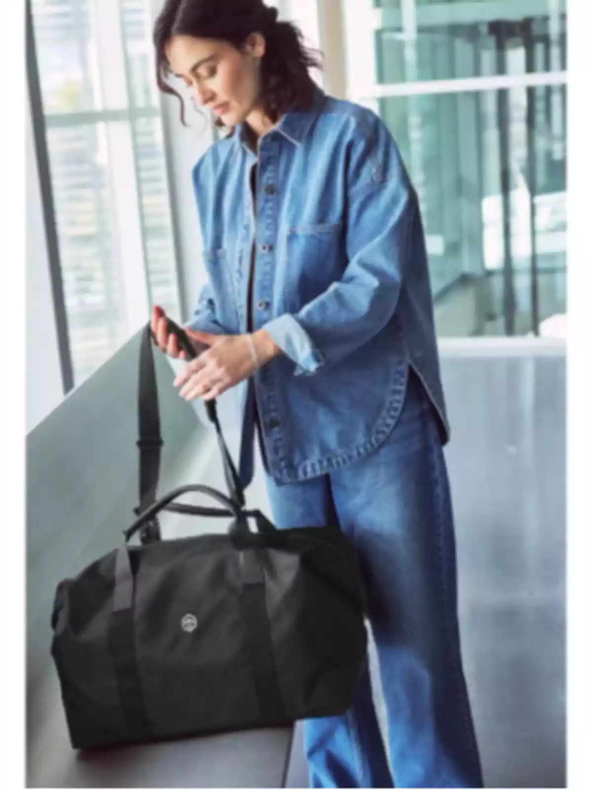 Dona amb camisa i pantalons texans sostenint una bossa de viatge negra en un entorn modern i lluminós Dona amb camisa i pantalons texans sostenint una bossa de viatge negra en un entorn modern i lluminós