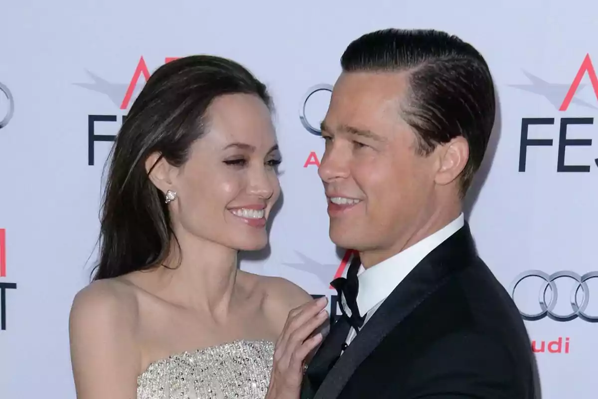Una pareja elegantemente vestida, que es Brad Pitt y Angelina Jolie, sonríe y posa junta en un evento con fondo de logotipos. Una pareja elegantemente vestida, que es Brad Pitt y Angelina Jolie, sonríe y posa junta en un evento con fondo de logotipos.
