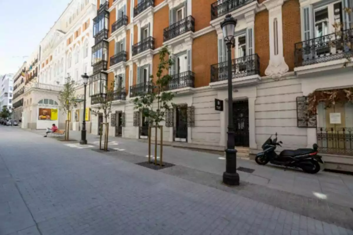 Carrer tranquil amb edificis de façana clàssica, arbres joves, una motocicleta aparcada i una persona asseguda en un banc. Carrer tranquil amb edificis de façana clàssica, arbres joves, una motocicleta aparcada i una persona asseguda en un banc.