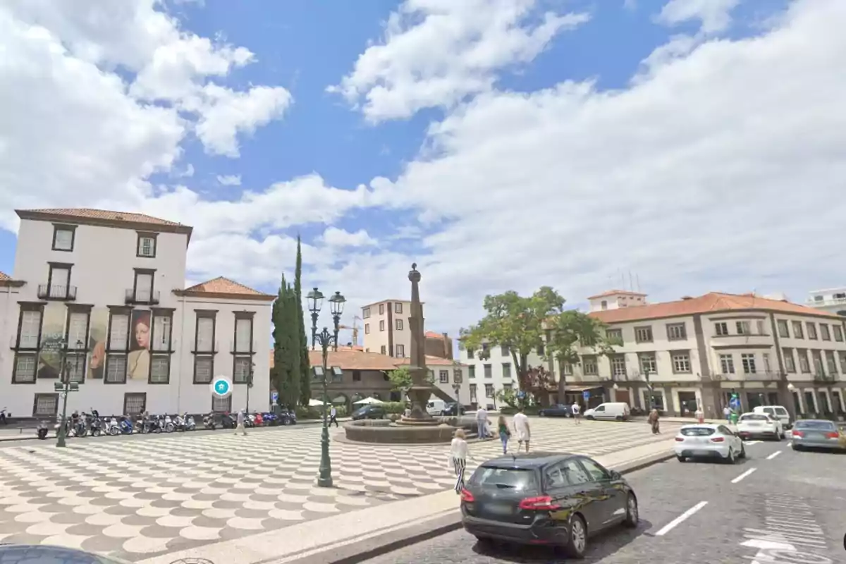 Plaça ampla amb una font central envoltada d’edificis històrics, cotxes aparcats i persones caminant sota un cel parcialment ennuvolat. Plaça ampla amb una font central envoltada d’edificis històrics, cotxes aparcats i persones caminant sota un cel parcialment ennuvolat.
