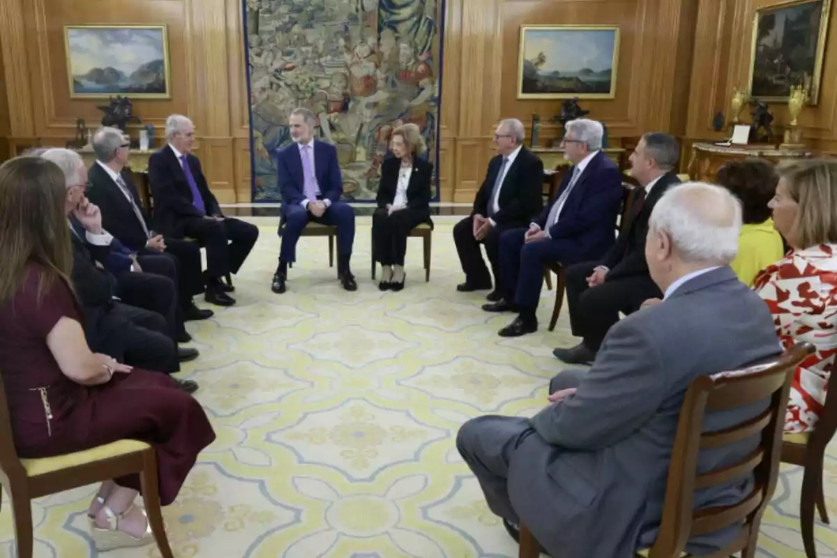Un grupo de personas elegantemente vestidas está sentado en círculo en una sala decorada con alfombra clara, tapiz y cuadros en las paredes