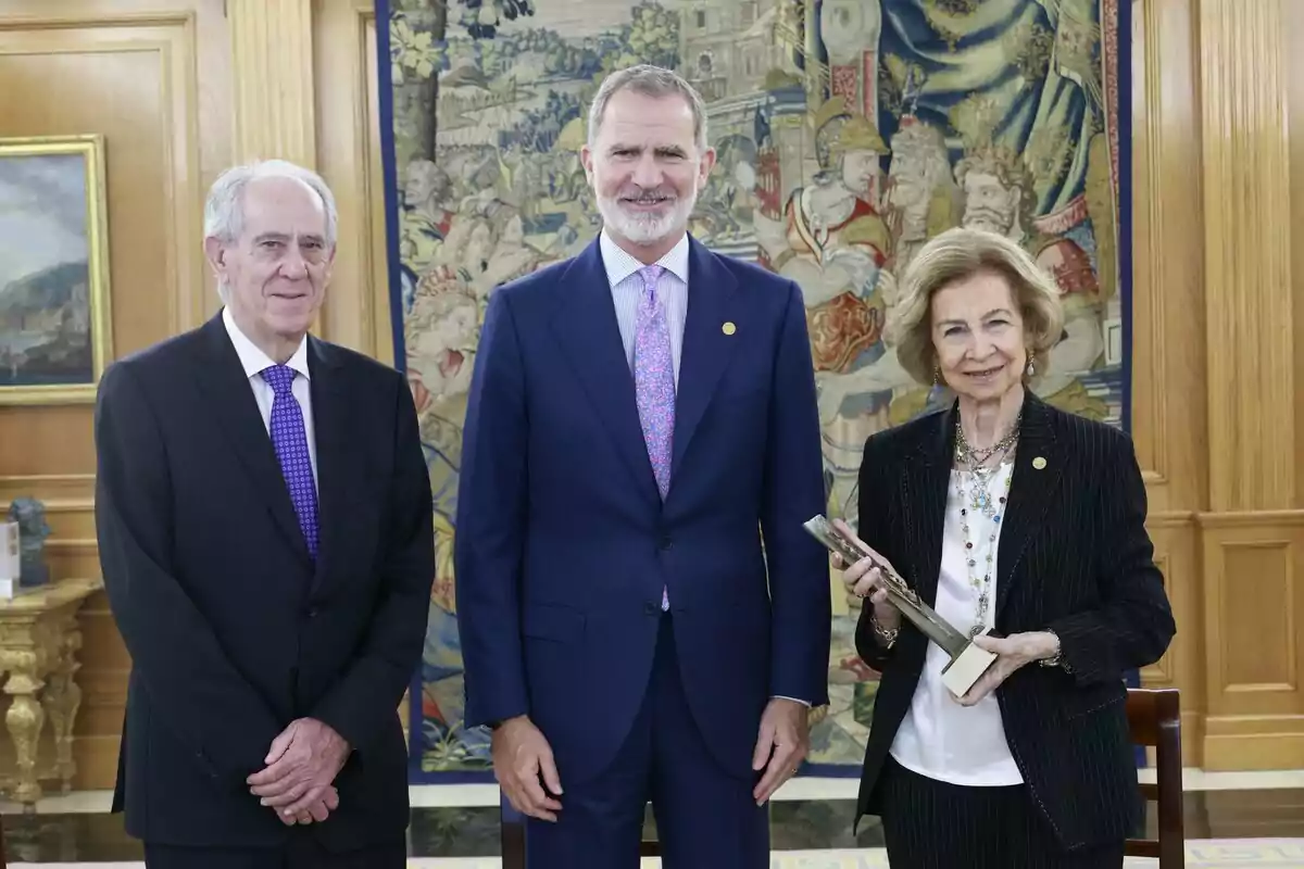 Felipe VI, la reina Sofía y otro hombre elegantemente vestidas posan en una sala decorada con un tapiz y muebles clásicos, una de ellas sostiene un trofeo rectangular plateado.
