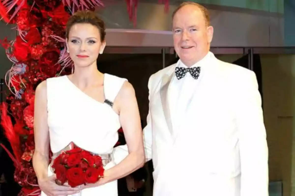 Pareja elegantemente vestida de blanco posando juntos, que son Alberto de Mónaco y Charlene, en un evento, ella sostiene un ramo de rosas rojas y hay una decoración floral roja al fondo. Pareja elegantemente vestida de blanco posando juntos, que son Alberto de Mónaco y Charlene, en un evento, ella sostiene un ramo de rosas rojas y hay una decoración floral roja al fondo.