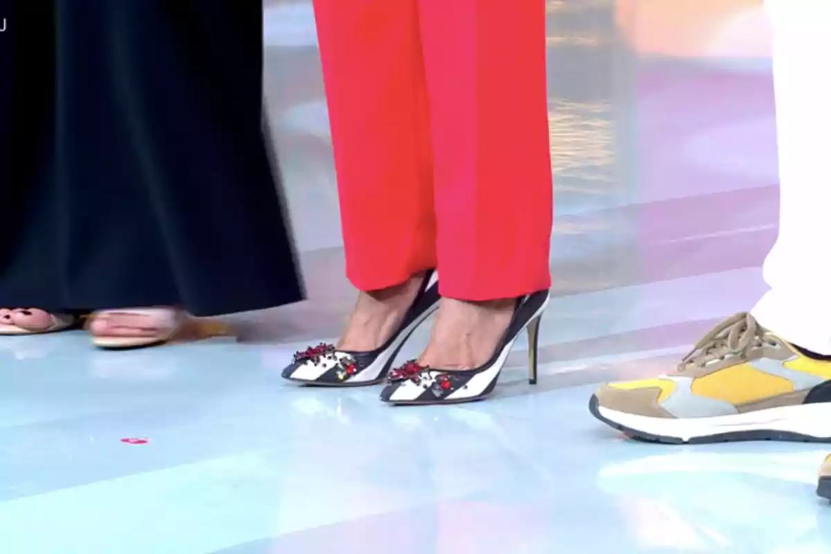 Tres personas de pie mostrando solo sus pies, una lleva sandalias, otra pantalón rojo con tacones de punta y la tercera zapatillas deportivas amarillas Tres personas de pie mostrando solo sus pies, una lleva sandalias, otra pantalón rojo con tacones de punta y la tercera zapatillas deportivas amarillas