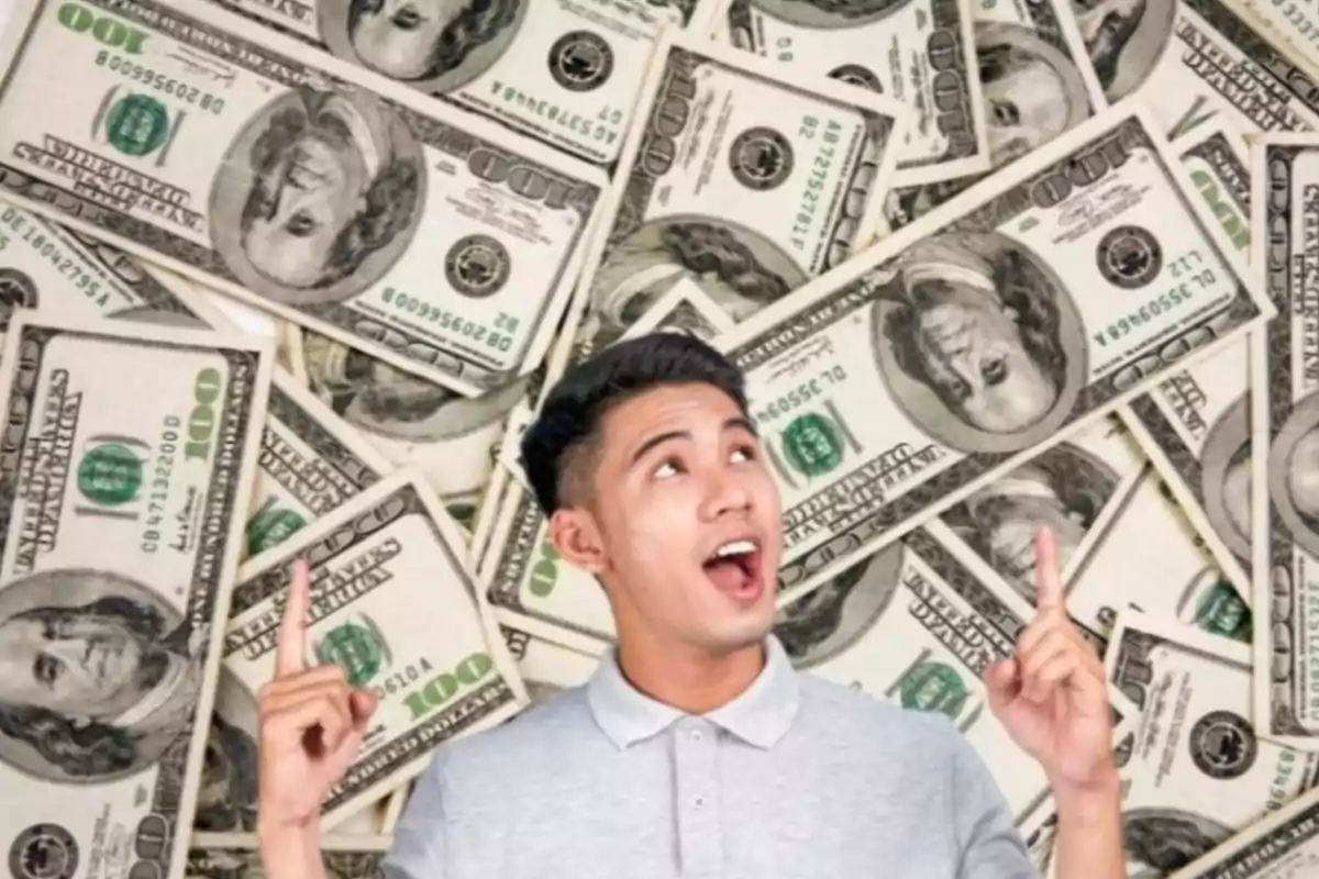 Smiling young man pointing upwards with both hands in front of a background of one hundred dollar bills Smiling young man pointing upwards with both hands in front of a background of one hundred dollar bills