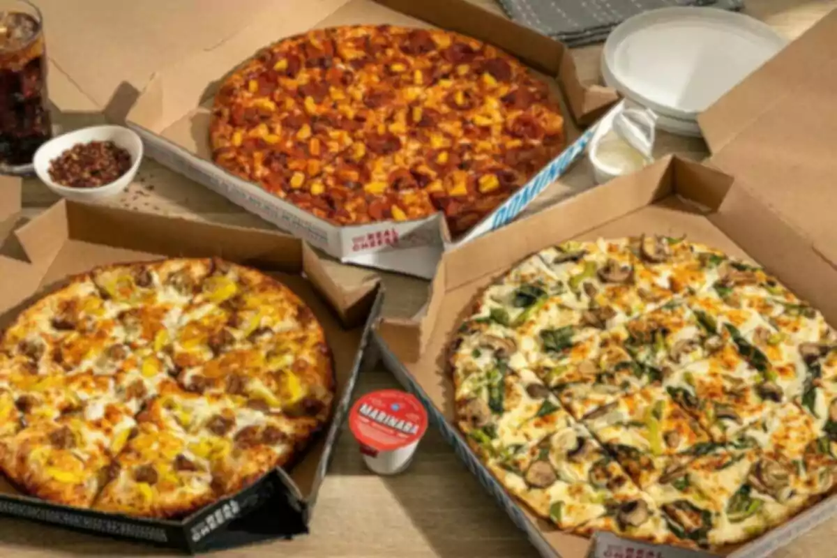 Tres pizzas diferentes en cajas de cartón sobre una mesa junto a un vaso de refresco, un recipiente con salsa marinara, un tazón pequeño con chile seco y platos blancos apilados Tres pizzas diferentes en cajas de cartón sobre una mesa junto a un vaso de refresco, un recipiente con salsa marinara, un tazón pequeño con chile seco y platos blancos apilados