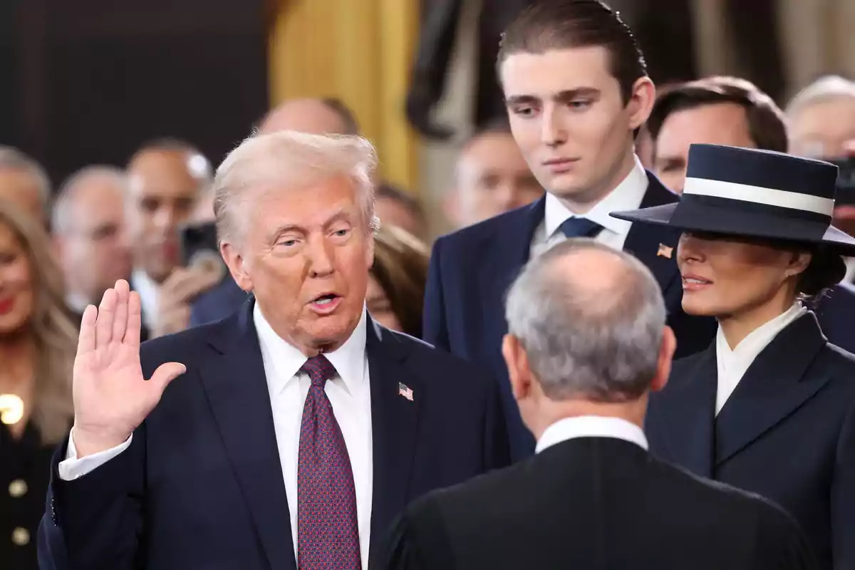 Donald Trump amb vestit fosc aixeca la mà dreta mentre és observat per diverses persones en un esdeveniment formal. Donald Trump amb vestit fosc aixeca la mà dreta mentre és observat per diverses persones en un esdeveniment formal.