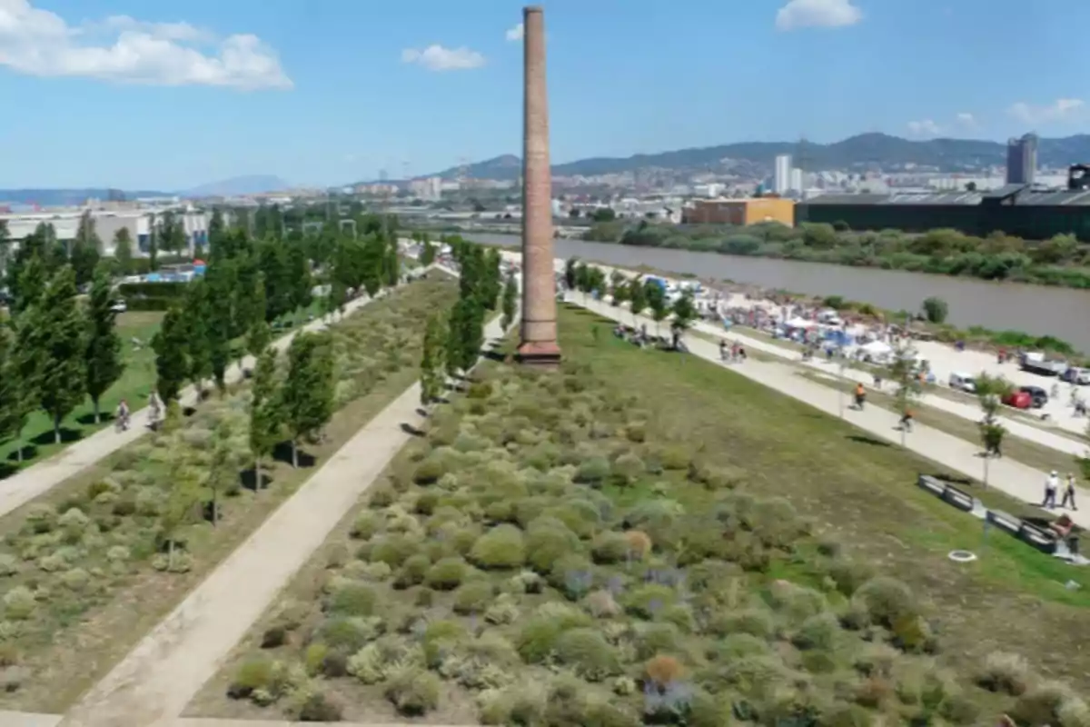 Vista d’un parc urbà amb camins de terra, vegetació i una xemeneia antiga al centre, al costat d’un riu i amb la ciutat al fons sota un cel serè. Vista d’un parc urbà amb camins de terra, vegetació i una xemeneia antiga al centre, al costat d’un riu i amb la ciutat al fons sota un cel serè.