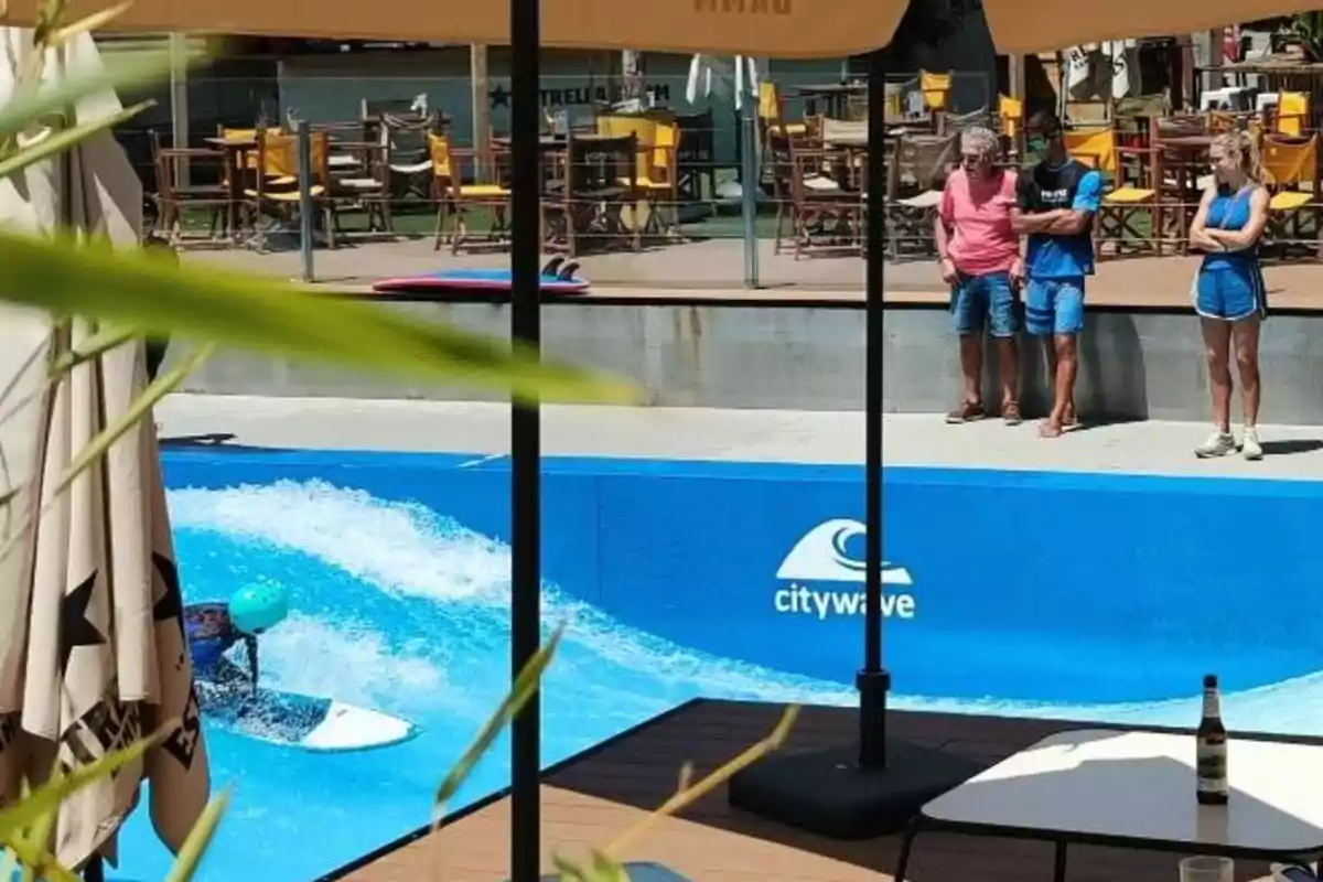 Persona surfeando en una piscina artificial mientras tres personas observan desde el borde y varias mesas y sillas están al fondo Persona surfeando en una piscina artificial mientras tres personas observan desde el borde y varias mesas y sillas están al fondo