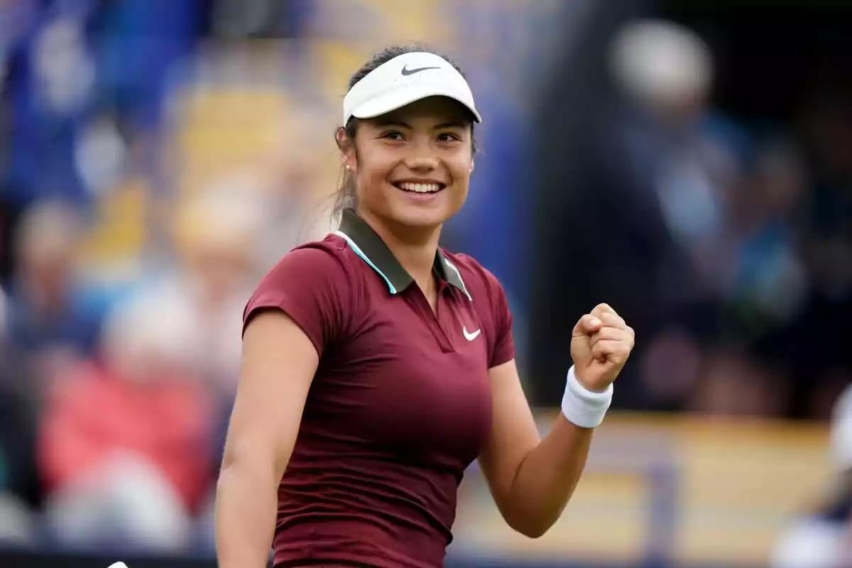 A smiling young tennis player, Emma Raducanu, celebrating on a court with a crowd in the background. A smiling young tennis player, Emma Raducanu, celebrating on a court with a crowd in the background.