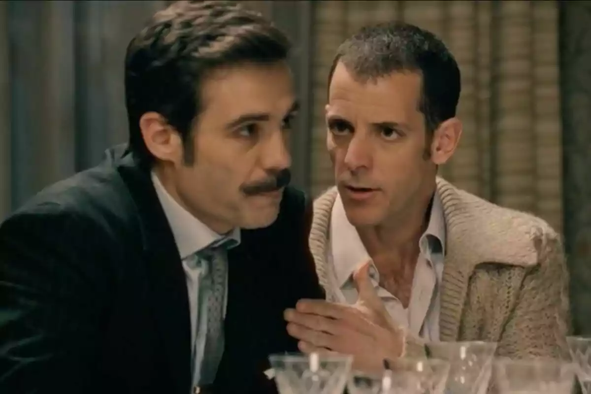 Two men are talking at a table with crystal glasses in front of them; one of them, Guillermo Barrientos, has a mustache and is wearing a suit, while the other is dressed in a beige sweater and appears to be speaking seriously. Two men are talking at a table with crystal glasses in front of them; one of them, Guillermo Barrientos, has a mustache and is wearing a suit, while the other is dressed in a beige sweater and appears to be speaking seriously.