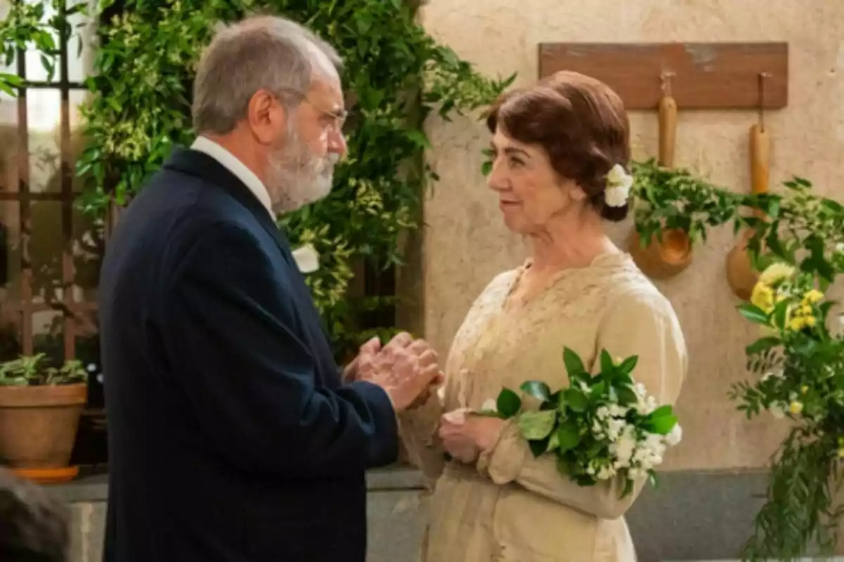 Parella de persones grans, que són Ròmul i Emília a 'La Promesa', vestides de manera elegant mirant-se amb afecte en un ambient decorat amb plantes i estris de cuina, la dona sosté un ram de flors blanques i verdes. Parella de persones grans, que són Ròmul i Emília a 'La Promesa', vestides de manera elegant mirant-se amb afecte en un ambient decorat amb plantes i estris de cuina, la dona sosté un ram de flors blanques i verdes.