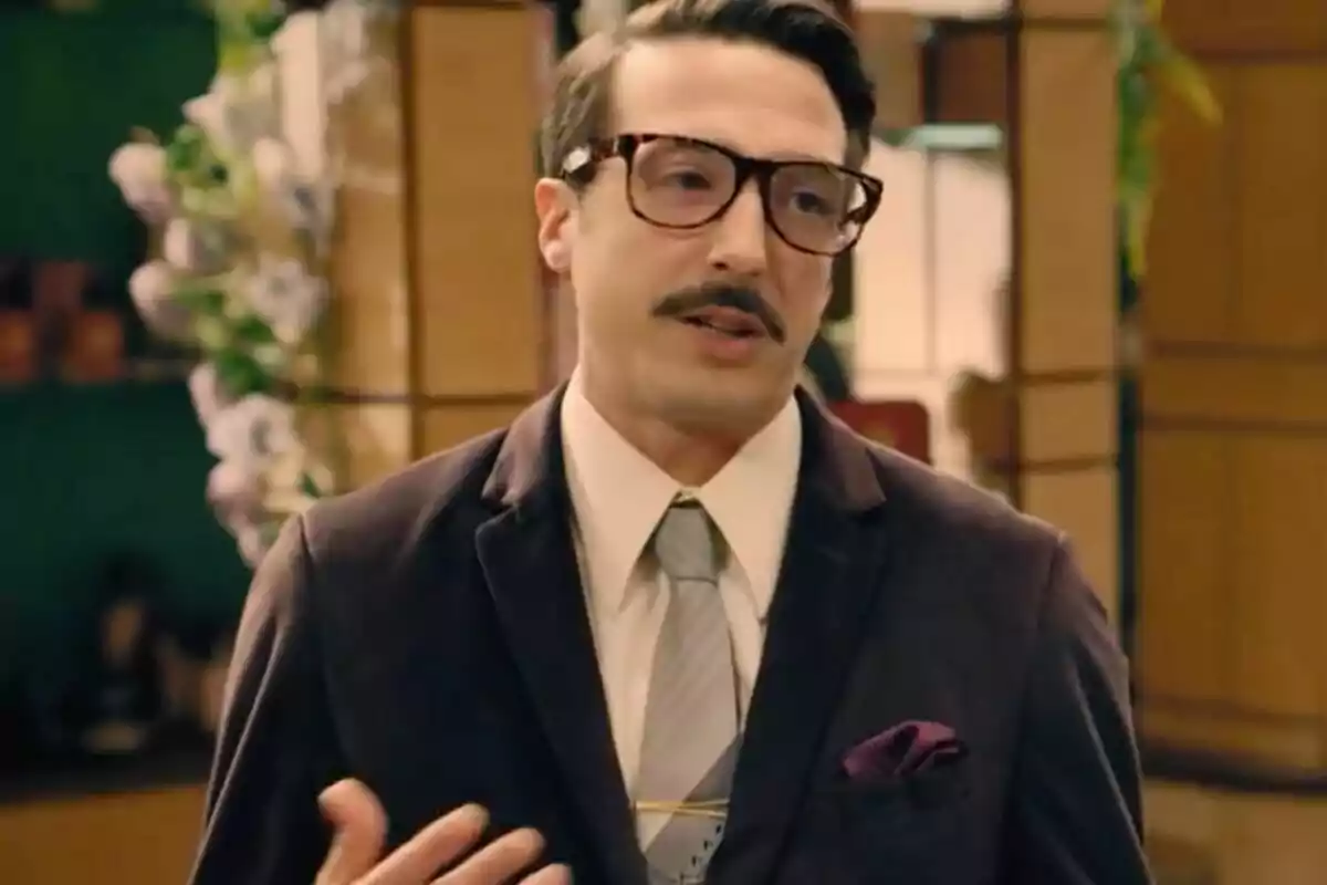 Cobeaga amb ulleres grosses i bigoti vestit amb un vestit fosc i corbata clara en un ambient interior elegant. Cobeaga amb ulleres grosses i bigoti vestit amb un vestit fosc i corbata clara en un ambient interior elegant.