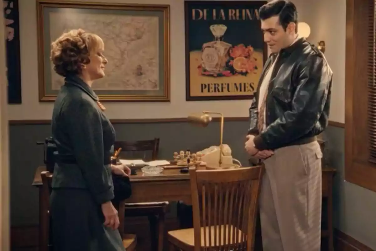 Una dona i un home, que són Irene i Tasío, conversen en una oficina decorada amb un mapa antic i un cartell de perfums, tots dos vestits amb roba d’època. Una dona i un home, que són Irene i Tasío, conversen en una oficina decorada amb un mapa antic i un cartell de perfums, tots dos vestits amb roba d’època.