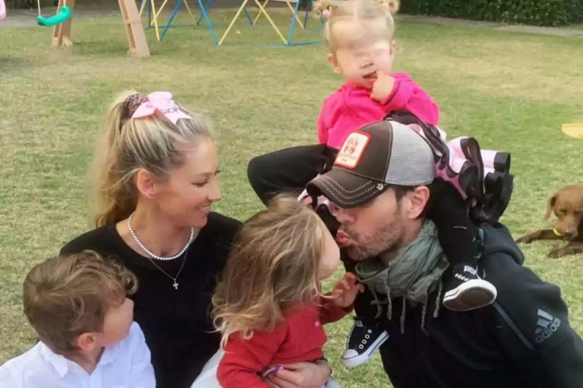 Familia Kournikova-Iglesias sentada en el césped disfrutando de un momento juntos mientras un hombre con gorra besa a una niña, una mujer sonríe y otros dos niños están cerca, al fondo se ve un perro acostado y juegos infantiles. Familia Kournikova-Iglesias sentada en el césped disfrutando de un momento juntos mientras un hombre con gorra besa a una niña, una mujer sonríe y otros dos niños están cerca, al fondo se ve un perro acostado y juegos infantiles.