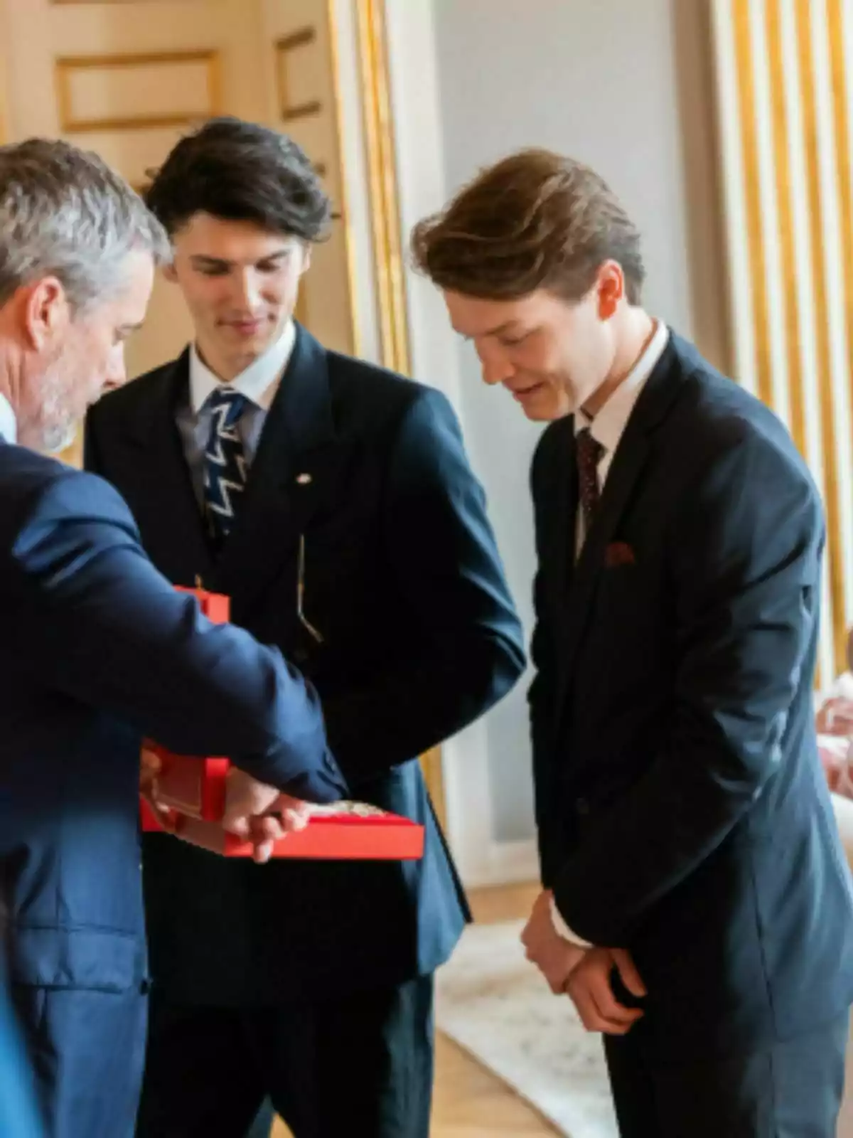 Tres hombres vestidos de traje participan en una ceremonia formal en un elegante salón, uno de ellos es Federico de Dinamarca y sostiene una caja roja que parece contener una medalla o reconocimiento. Tres hombres vestidos de traje participan en una ceremonia formal en un elegante salón, uno de ellos es Federico de Dinamarca y sostiene una caja roja que parece contener una medalla o reconocimiento.