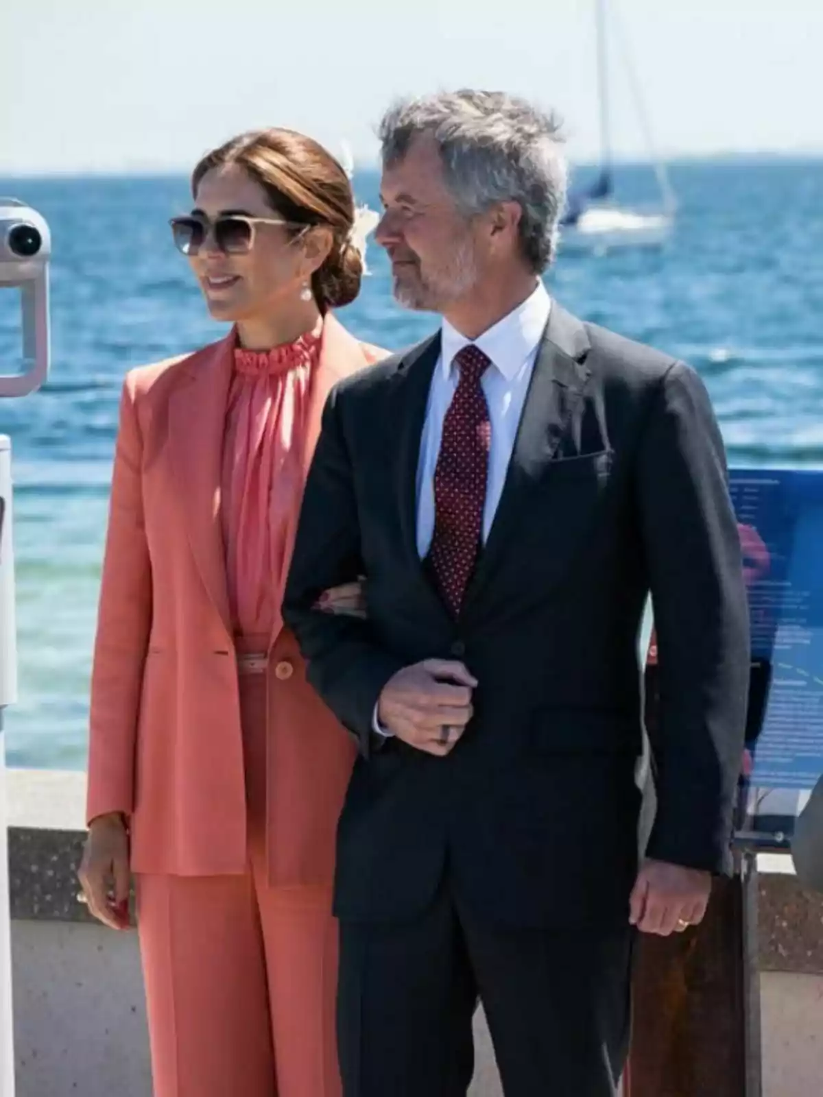 An elegantly dressed couple, Federico and Mary of Denmark, pose smiling by the sea on a sunny day. An elegantly dressed couple, Federico and Mary of Denmark, pose smiling by the sea on a sunny day.