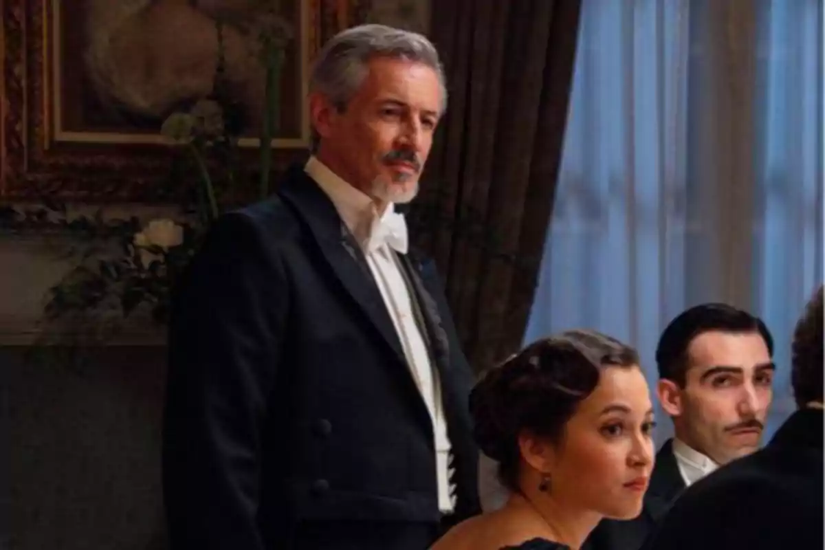 Tres personas elegantemente vestidas están sentadas alrededor de una mesa en una cena formal, con un hombre de cabello canoso de pie al fondo Tres personas elegantemente vestidas están sentadas alrededor de una mesa en una cena formal, con un hombre de cabello canoso de pie al fondo
