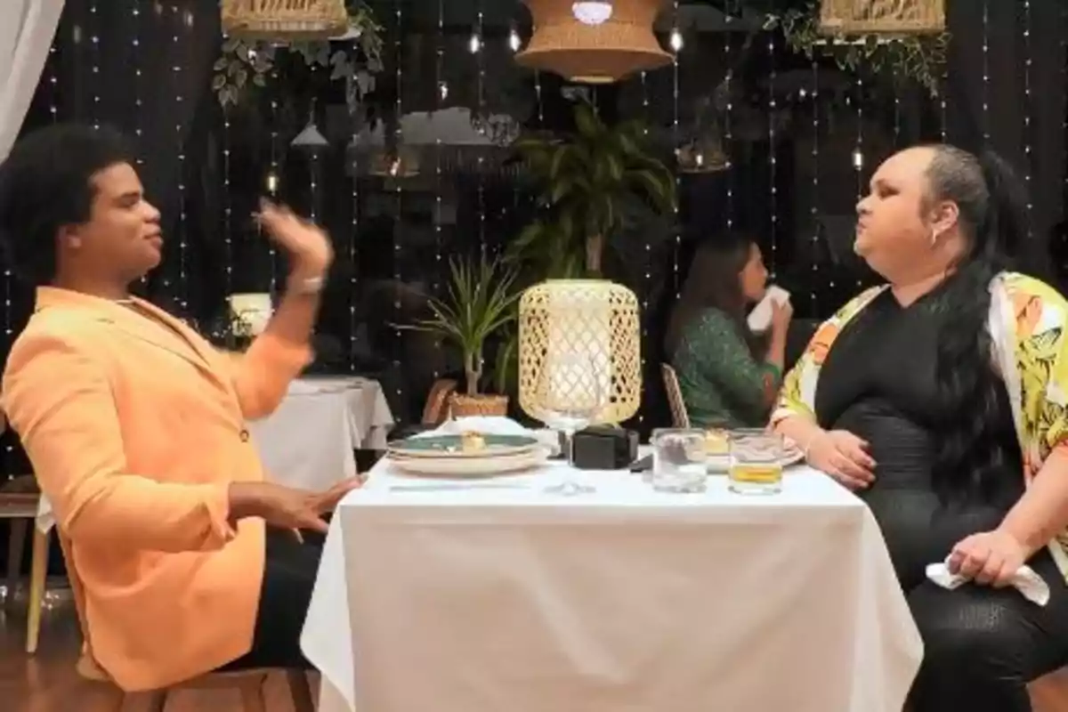 Dues persones assegudes cara a cara en una taula d’un restaurant elegant, una d’elles gesticulant amb la mà mentre l’altra observa amb expressió seriosa, amb decoració de llums i plantes al fons. Captura de ‘First Dates’. Dues persones assegudes cara a cara en una taula d’un restaurant elegant, una d’elles gesticulant amb la mà mentre l’altra observa amb expressió seriosa, amb decoració de llums i plantes al fons. Captura de ‘First Dates’.