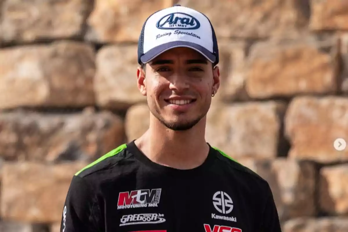 Borja Gómez con gorra blanca y azul y camiseta negra con logotipos de motociclismo sonriendo frente a una pared de piedra Borja Gómez con gorra blanca y azul y camiseta negra con logotipos de motociclismo sonriendo frente a una pared de piedra