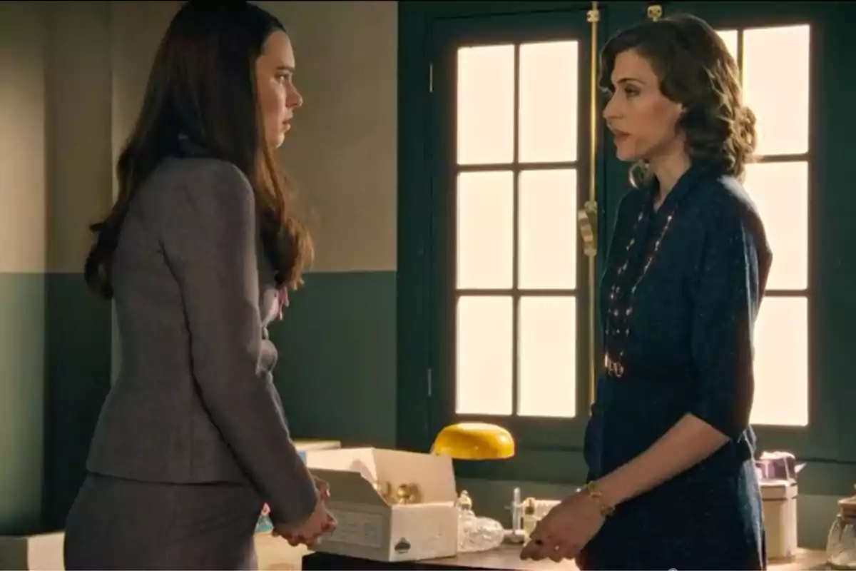 Marta de la Reina (Marta Belmonte) i Fina Valero (Alba Brunet) conversen dretes en una oficina il·luminada per la llum que entra per una finestra, una d’elles porta un vestit gris i l’altra un vestit blau fosc. Marta de la Reina (Marta Belmonte) i Fina Valero (Alba Brunet) conversen dretes en una oficina il·luminada per la llum que entra per una finestra, una d’elles porta un vestit gris i l’altra un vestit blau fosc.