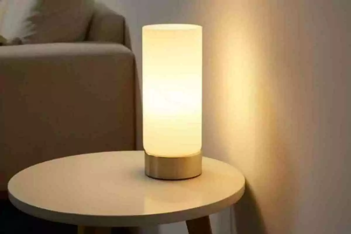 Lámpara de mesa cilíndrica encendida sobre una mesa redonda junto a un sofá