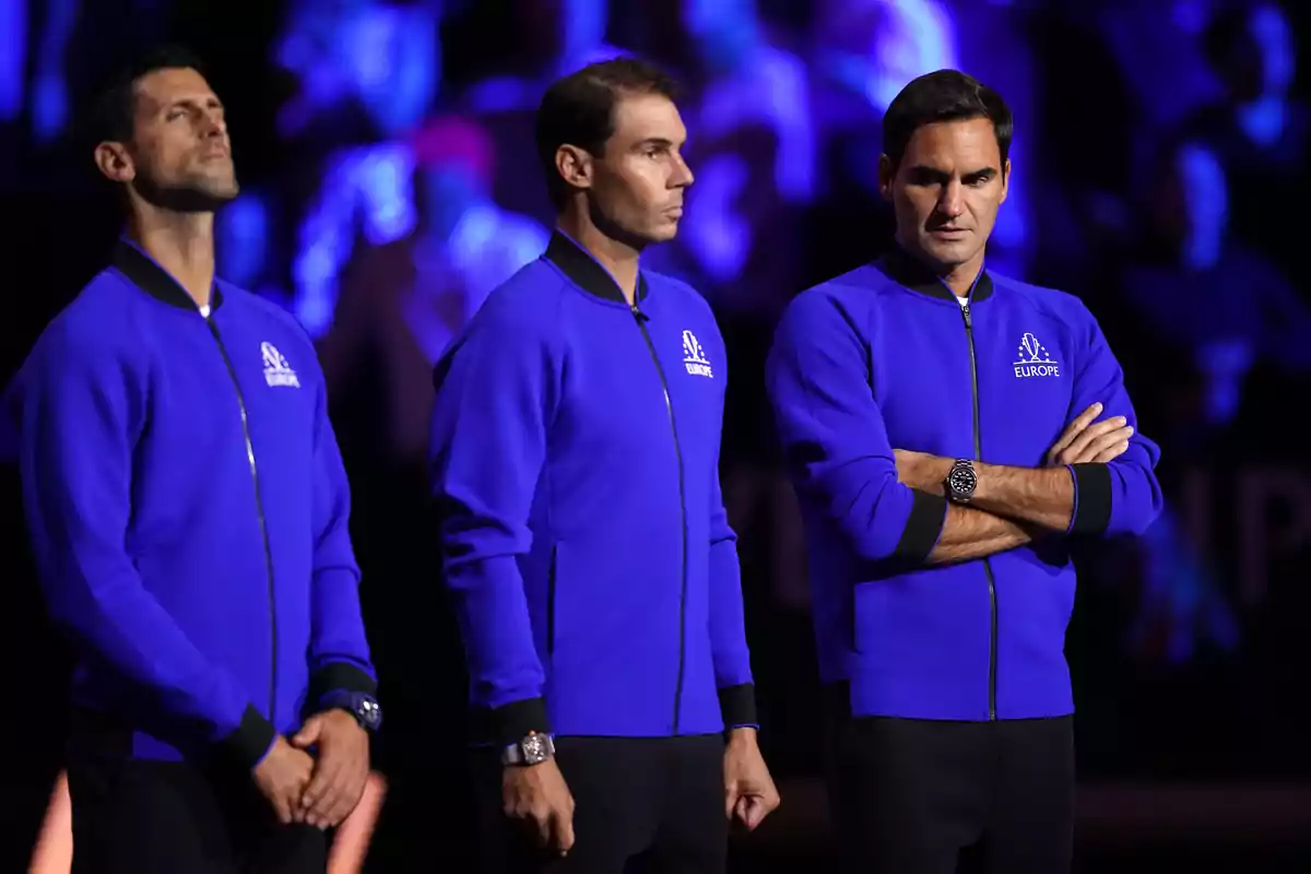 Rafa Nadal, Roger Federer i Novak Djokovic amb jaquetes blaves de l’equip Europa estan drets en un escenari il·luminat amb llums morades i blaves, mirant cap endavant amb expressions serioses. Rafa Nadal, Roger Federer i Novak Djokovic amb jaquetes blaves de l’equip Europa estan drets en un escenari il·luminat amb llums morades i blaves, mirant cap endavant amb expressions serioses.
