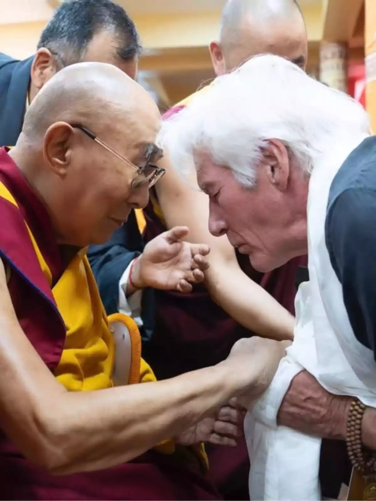 Ricard Gere with white hair bows his head in respect while holding the hands of a Buddhist monk dressed in yellow and red robes. Ricard Gere with white hair bows his head in respect while holding the hands of a Buddhist monk dressed in yellow and red robes.