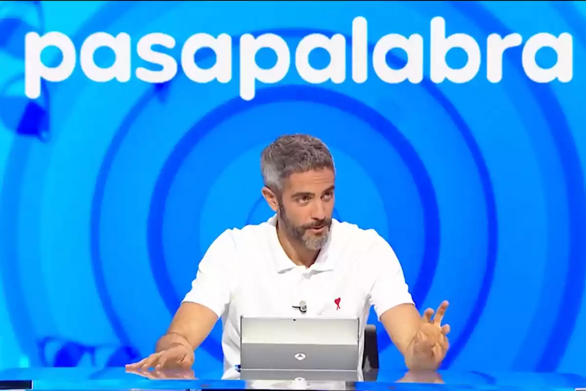 Roberto Leal de cabells canosos i barba assegut davant d’una taula amb una tauleta, al fons el logotip de Pasapalabra sobre un fons blau Roberto Leal de cabells canosos i barba assegut davant d’una taula amb una tauleta, al fons el logotip de Pasapalabra sobre un fons blau