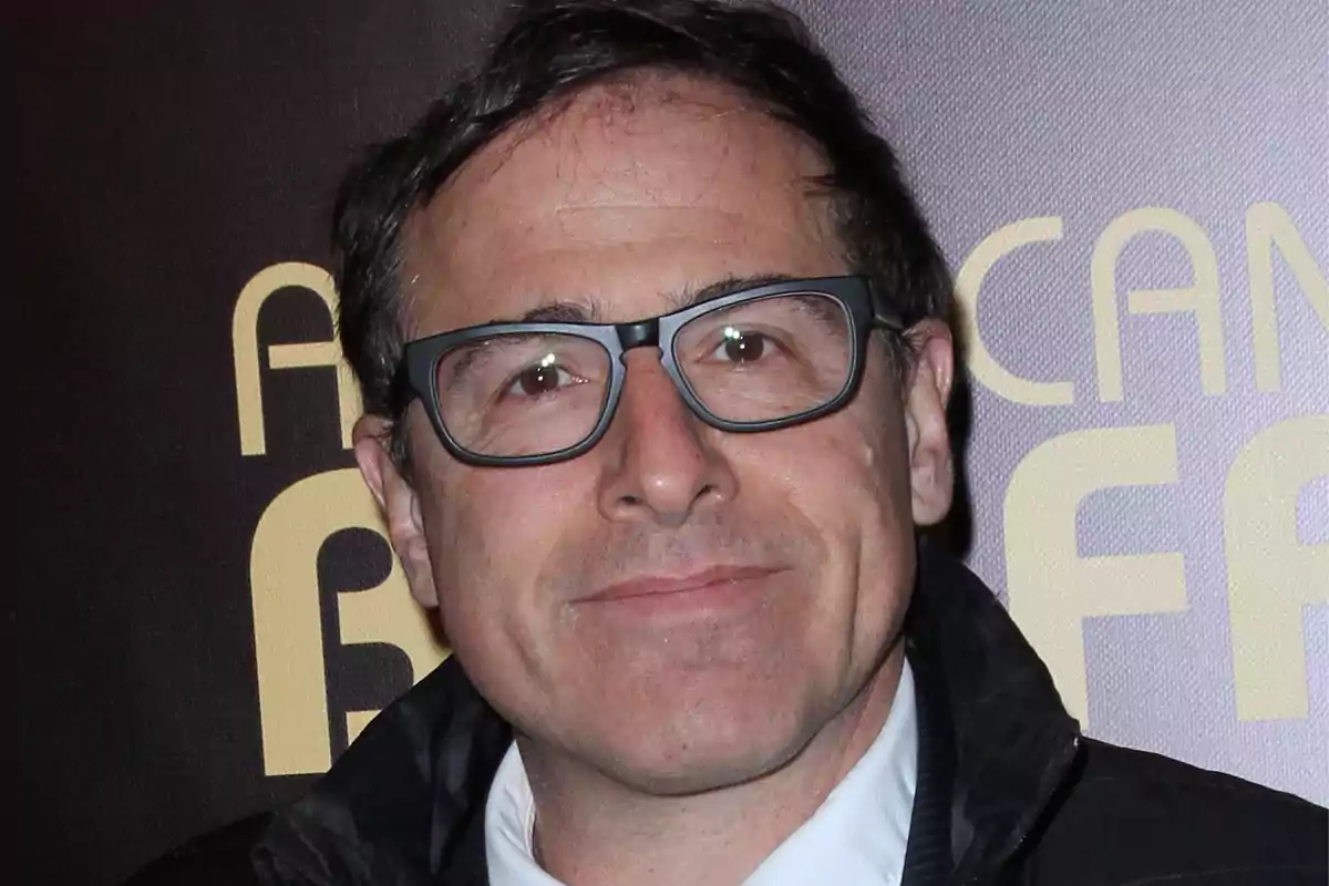 David O. Russell de cabello corto y gafas oscuras sonriendo levemente frente a un fondo con letras grandes desenfocadas. David O. Russell de cabello corto y gafas oscuras sonriendo levemente frente a un fondo con letras grandes desenfocadas.