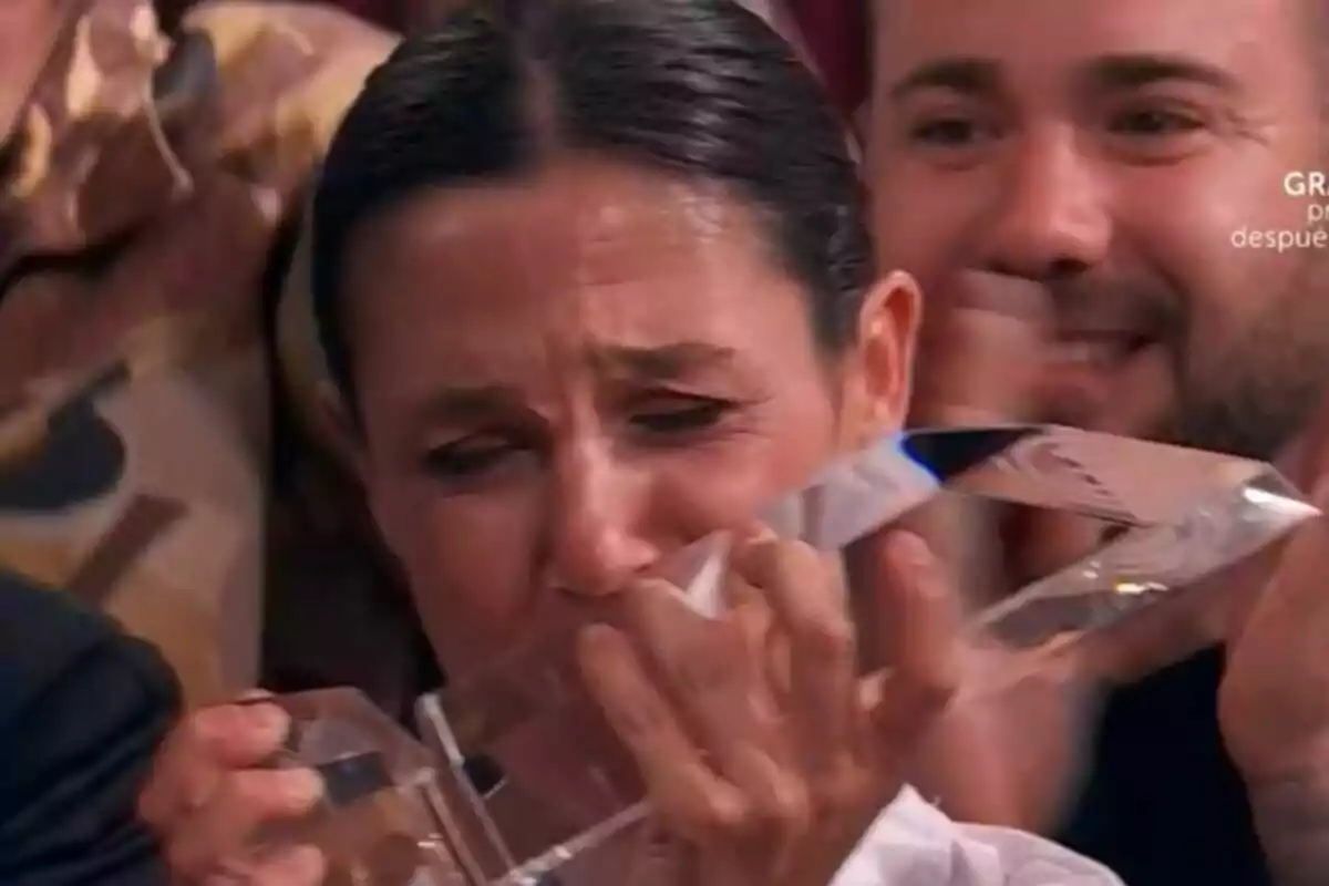 Gabriela emocionada sosté un trofeu de vidre de MasterChef mentre plora d’alegria envoltada de persones que la donen suport. Gabriela emocionada sosté un trofeu de vidre de MasterChef mentre plora d’alegria envoltada de persones que la donen suport.