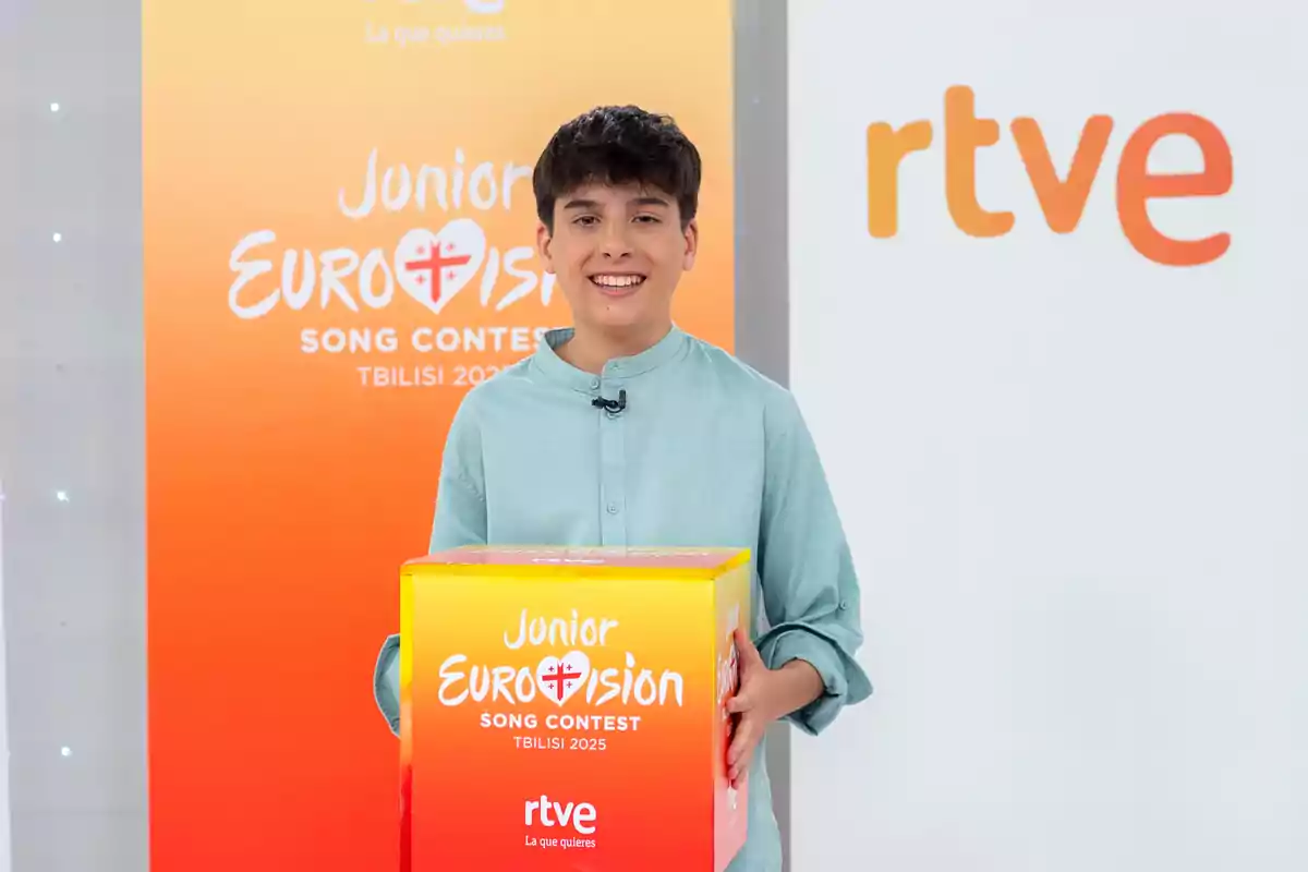 Un jove somrient sosté una caixa del Junior Eurovision Song Contest Tbilisi 2025 davant d’un fons amb els logotips de RTVE i el concurs. Un jove somrient sosté una caixa del Junior Eurovision Song Contest Tbilisi 2025 davant d’un fons amb els logotips de RTVE i el concurs.