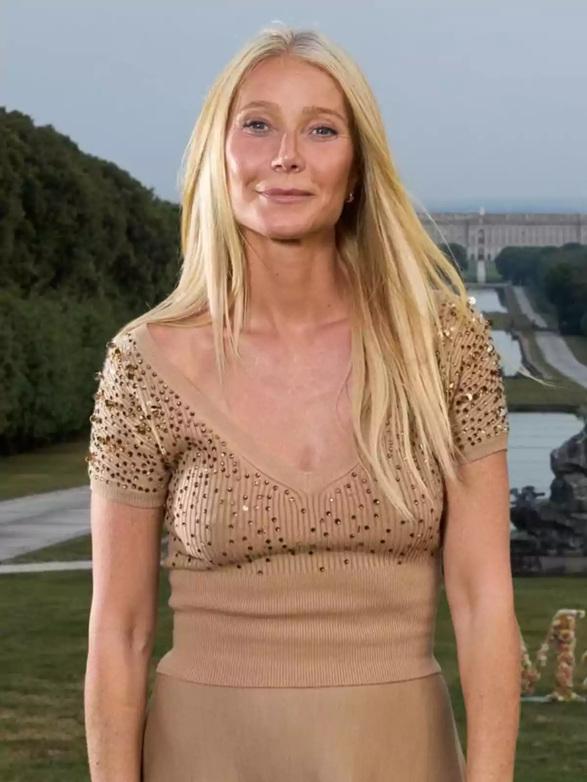 Gwyneth Paltrow amb vestit beix decorat amb pedreria posant a l’aire lliure davant d’un jardí i una font Gwyneth Paltrow amb vestit beix decorat amb pedreria posant a l’aire lliure davant d’un jardí i una font