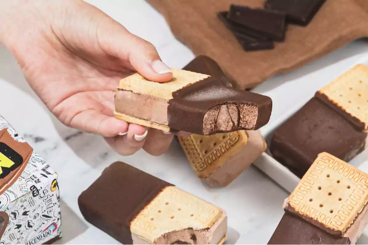 Una mà sosté un entrepà de gelat de xocolata parcialment cobert de xocolata, envoltat d’altres entrepans similars sobre una taula. Una mà sosté un entrepà de gelat de xocolata parcialment cobert de xocolata, envoltat d’altres entrepans similars sobre una taula.