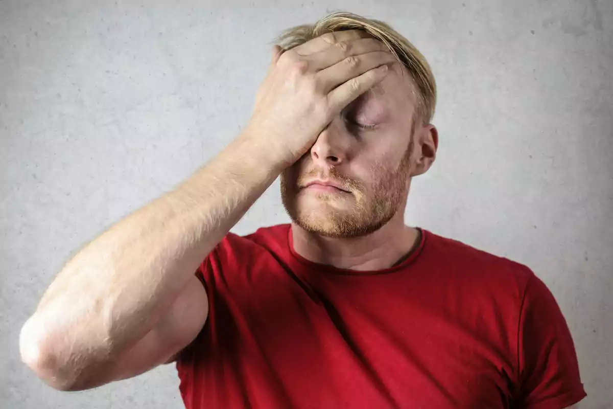Man wearing a red t-jersey covering his face with one hand, showing an expression of frustration Man wearing a red t-jersey covering his face with one hand, showing an expression of frustration
