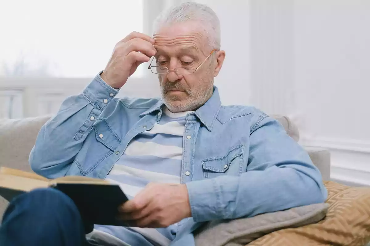 Older man sitting on a sofa reading a book and wearing glasses while looking thoughtful Older man sitting on a sofa reading a book and wearing glasses while looking thoughtful