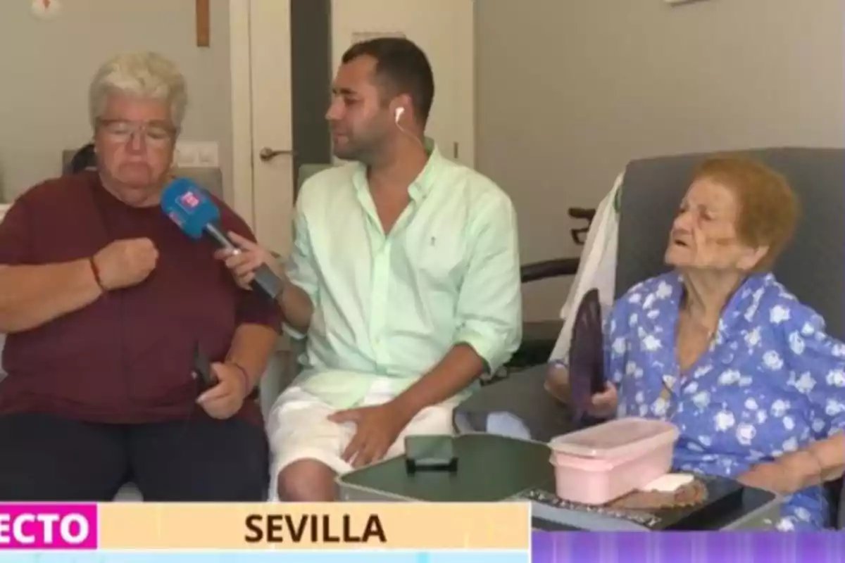 Un reportero entrevista a dos mujeres mayores en una sala, una de ellas sostiene un abanico y la otra parece emocionada, en la parte inferior aparece el texto Sevilla. Un reportero entrevista a dos mujeres mayores en una sala, una de ellas sostiene un abanico y la otra parece emocionada, en la parte inferior aparece el texto Sevilla.