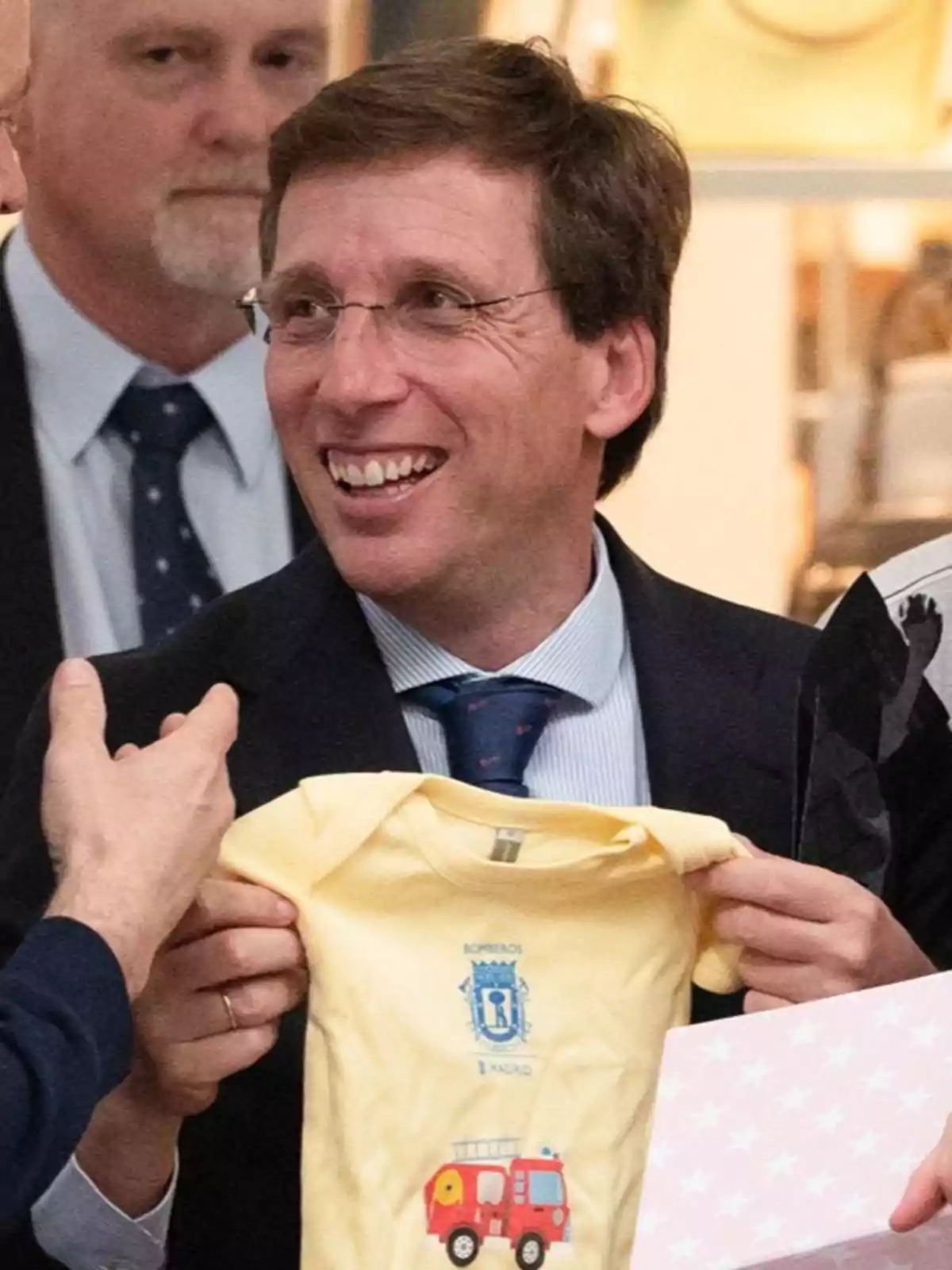 Almeida sonriente con gafas sostiene un body amarillo de bebé con un dibujo de camión de bomberos y el escudo de Madrid Almeida sonriente con gafas sostiene un body amarillo de bebé con un dibujo de camión de bomberos y el escudo de Madrid