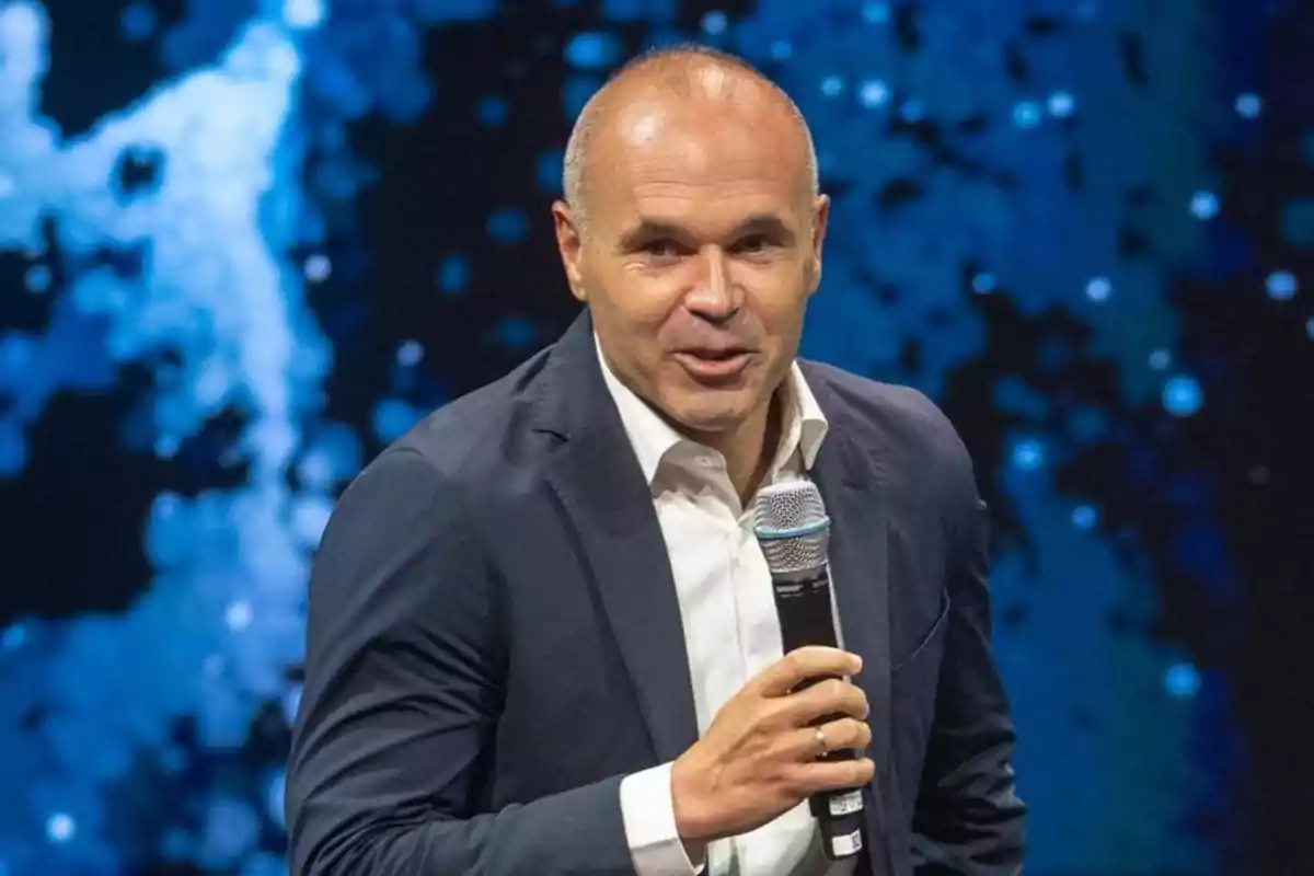 Andrés Iniesta calvo con saco oscuro y camisa blanca hablando al micrófono en un escenario con fondo azul desenfocado Andrés Iniesta calvo con saco oscuro y camisa blanca hablando al micrófono en un escenario con fondo azul desenfocado