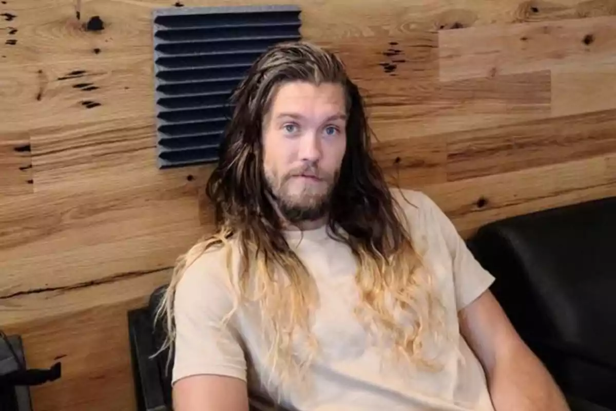 Bryan Braman de cabello largo y barba sentado en un sillón frente a una pared de madera Bryan Braman de cabello largo y barba sentado en un sillón frente a una pared de madera