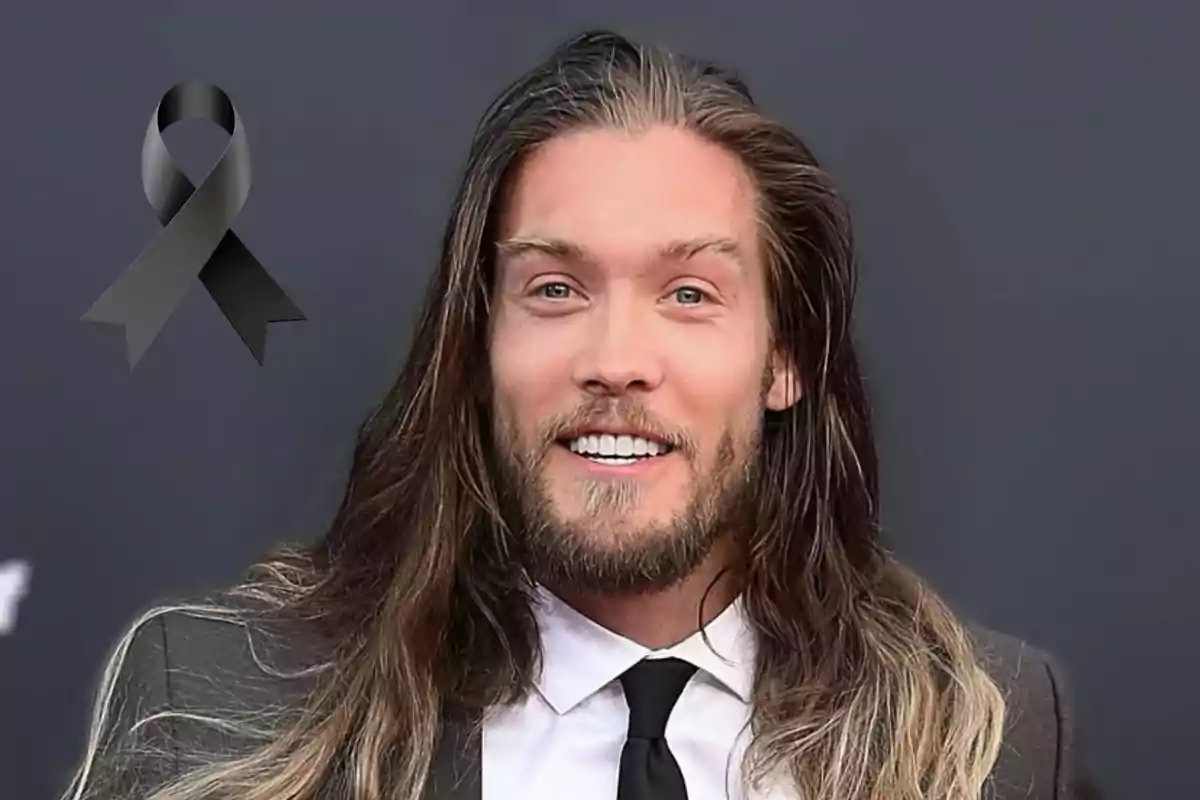 Bryan Braman de cabello largo y barba con traje y corbata sonriendo frente a un fondo oscuro con un moño negro en señal de luto en la esquina superior izquierda