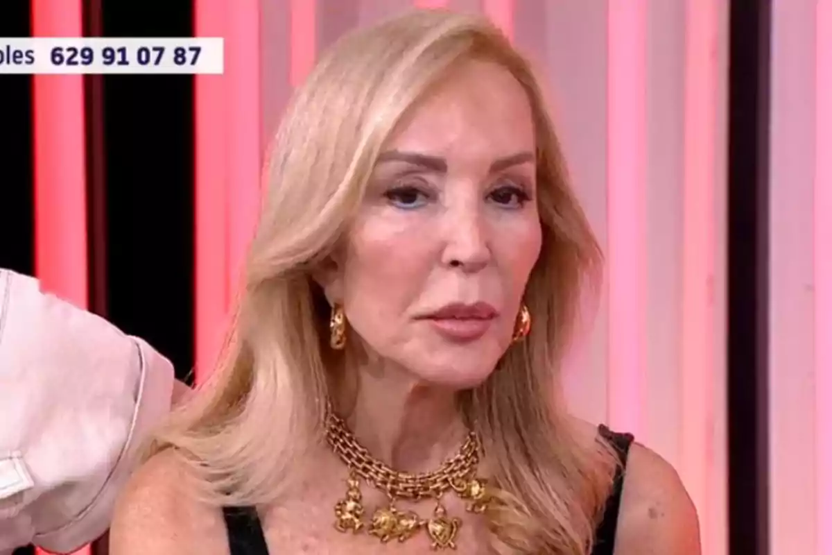 Carmen Lomana rubia de cabello largo con collar y aretes dorados sentada en un set de televisión con luces rosadas de fondo Carmen Lomana rubia de cabello largo con collar y aretes dorados sentada en un set de televisión con luces rosadas de fondo