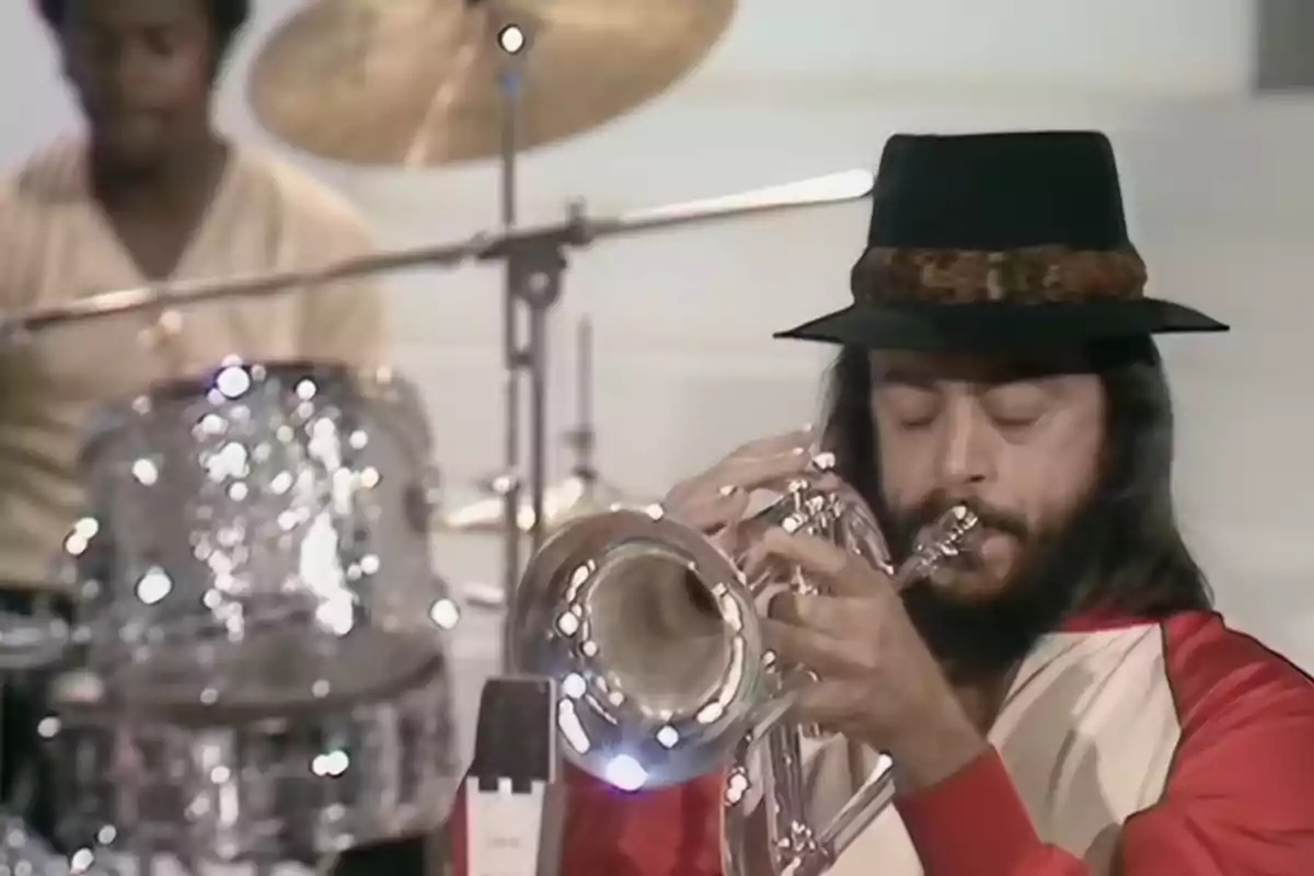Chuck Mangione con sombrero negro y barba tocando un instrumento de viento metálico mientras un baterista toca al fondo