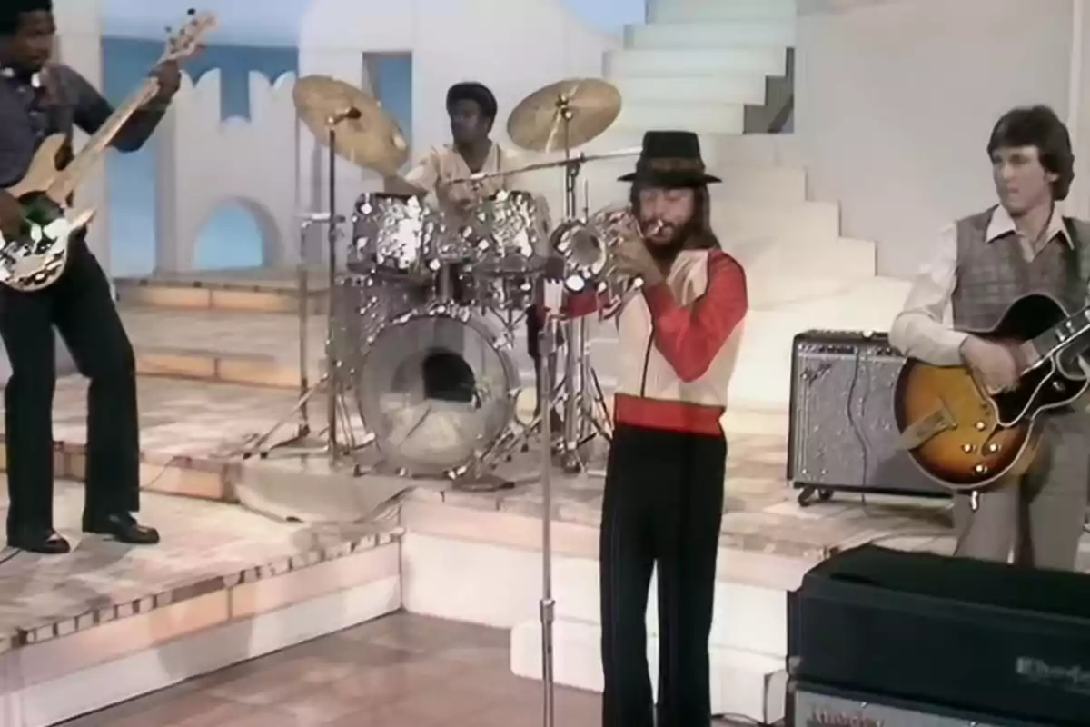 Cuatro músicos tocan instrumentos en un escenario con fondo blanco y escaleras uno toca el bajo otro la batería otro la trompeta y otro la guitarra eléctrica