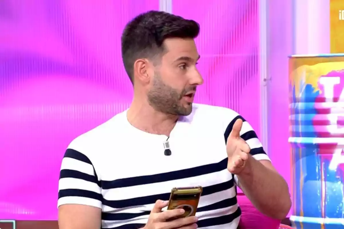 Hombre con barba y camiseta de rayas blancas y negras sostiene un teléfono móvil mientras gesticula en un set de televisión con fondo rosa
