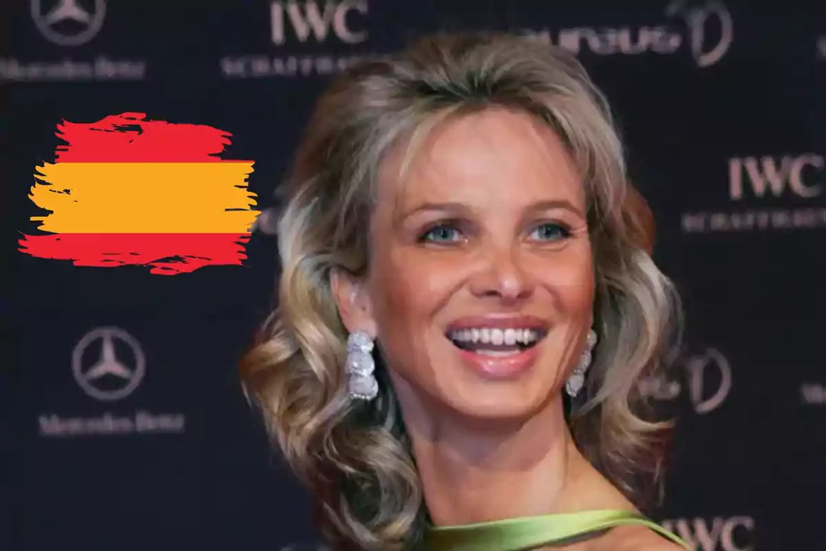 Corinna Larsen rubia sonriente con pendientes grandes en un evento elegante y una bandera de España pintada al fondo