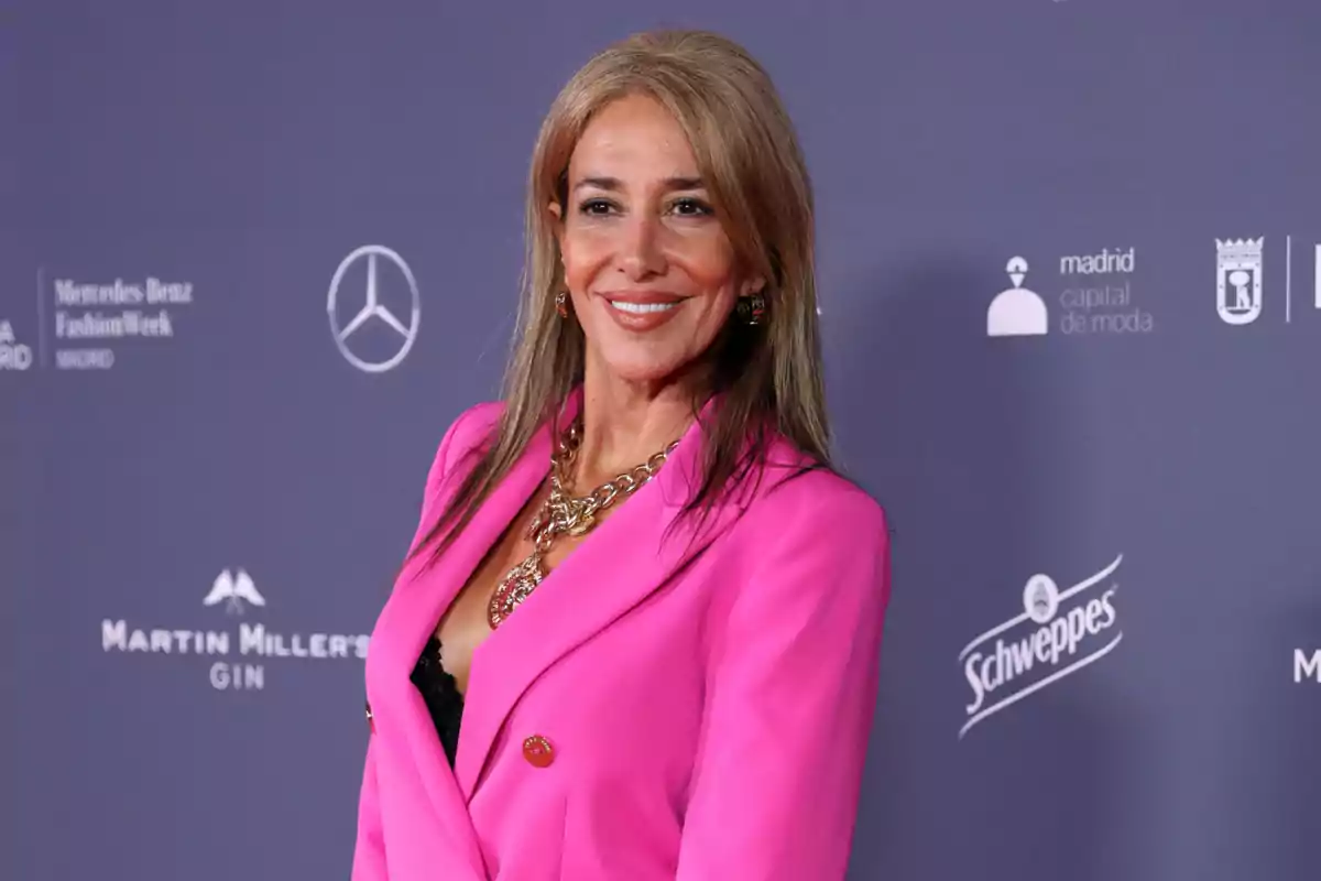 Mujer sonriendo con un saco rosa brillante y collar dorado posando frente a un fondo con logotipos de marcas en un evento de moda